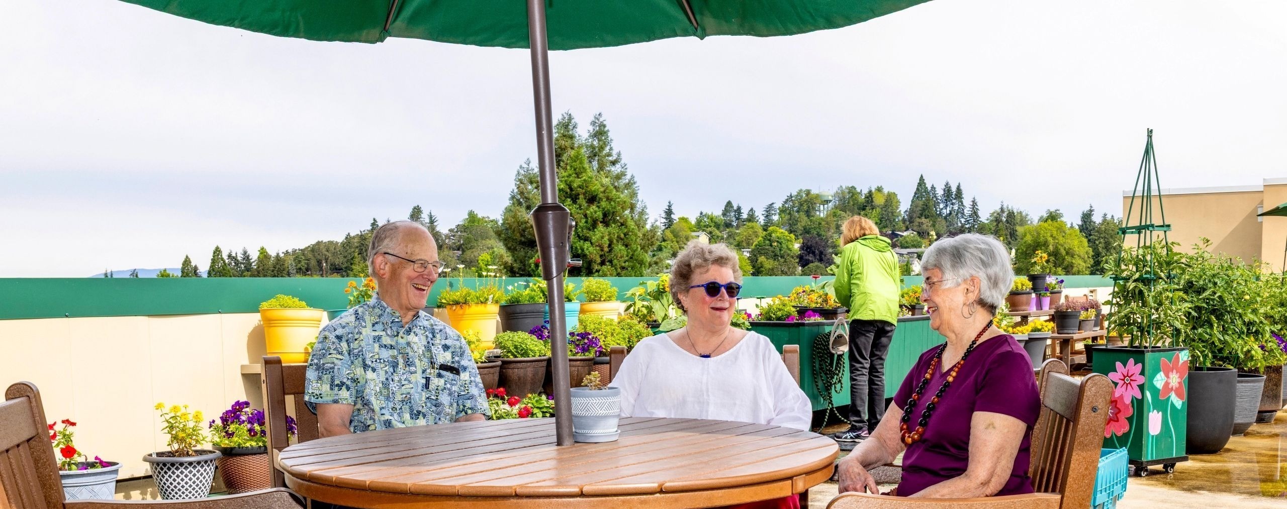 Three smiling older adults sit at a table on a rooftop garden, surrounded by potted plants. Another person tends to plants in the background.