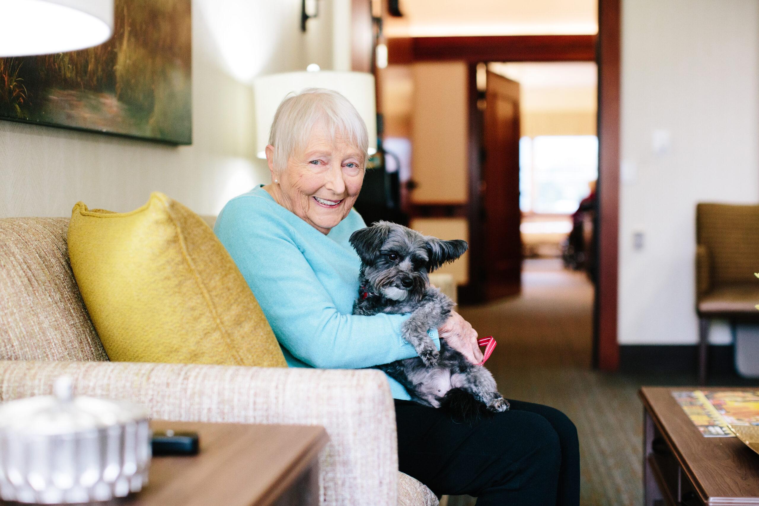 An older woman sitting in a chair smiles as she holds a dog