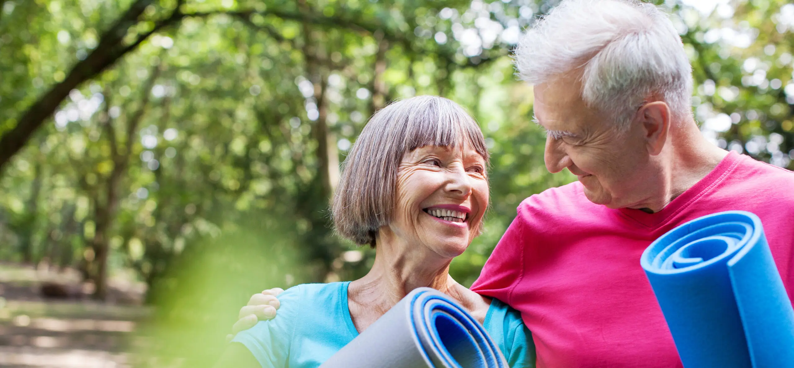 A smiling couple looks at each other and holds yoga mats in the park